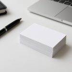 A stack of blank business cards next to a pen and a laptop on a desk.